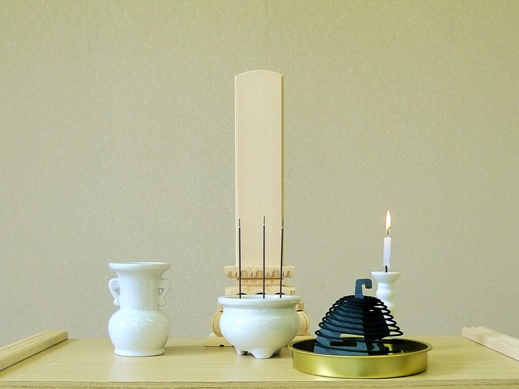 白い花立て、３本のお線香が立てられた白い香炉、白い燭台、渦巻き線香の後ろにある白木位牌