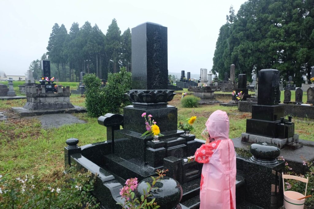 雨の降る中でお墓に手を合わせるピンク色のレインコートを着た少女